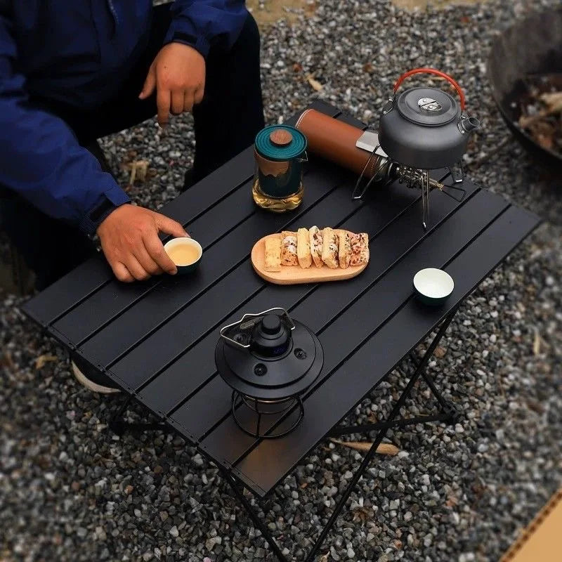 Roll-Up Camping Table overhead campsite view with a camper reaching for a cup beside snacks and a kettle.