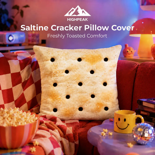A novelty throw pillow designed to look like a toasted saltine cracker, styled on a red couch in a cozy movie-night setting with popcorn, a yellow smiley face mug, and ambient blue and red lighting. The HighPeak brand logo and text are visible at the top.