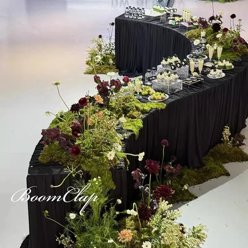 Serpentine Event Display Table draped in black linens with refreshments, green foliage and dark red flowers.
