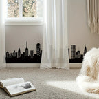 Skyline Wall Decal applied along a bedroom baseboard behind sheer curtains in a bright reading corner