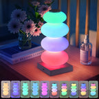 Stacked Pebble RGB Lamp glowing green, blue, purple and pink beside flowers and books.