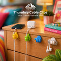 Thumbs up cable clips in yellow, green, blue, and cloud-shaped white holders mounted on a wooden nightstand, holding a black smartphone, a white wireless earbud case, and white wired earbuds. The background is a cozy, softly blurred bedroom setting with a lamp, books, and pillows.