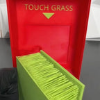 Touch Grass Meme Box with a gloved hand lifting the green grass insert from the red interior