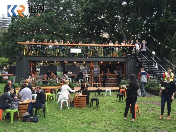 Wide view of a black two-story container restaurant at an outdoor event with guests on both levels