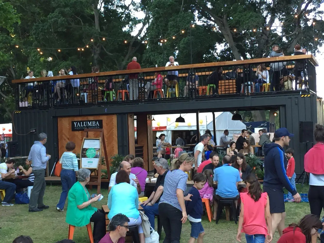 Black two-story container restaurant operating as a busy festival venue with rooftop and ground seating