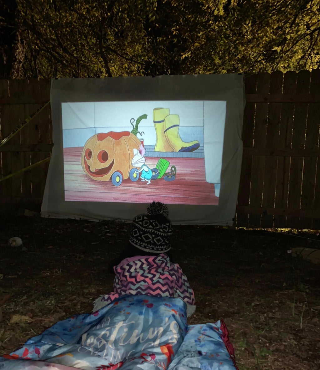 HighPeak MiniProjector™ projecting a colorful cartoon on an outdoor screen with a child watching in a cozy blanket at night