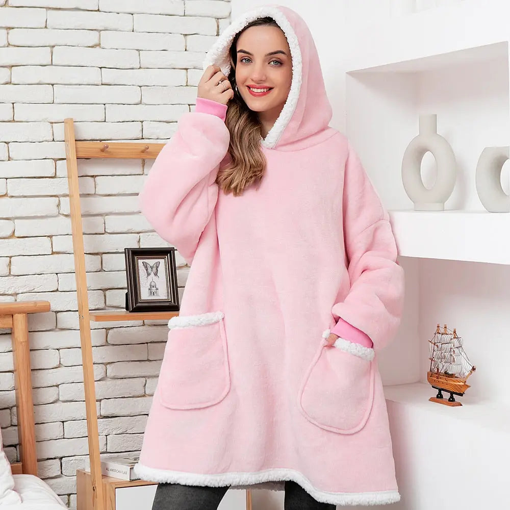Pink oversized hoodie blanket with sherpa lining and front pockets shown against white brick wall in cozy home setting