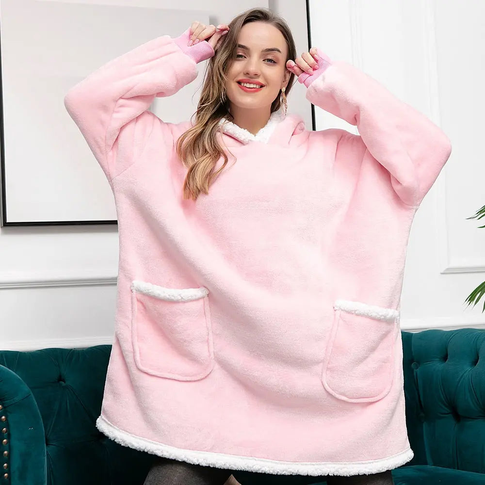 Oversized Hoodie Blanket in soft pink with sherpa lining and front pockets, shown on sofa against white wall