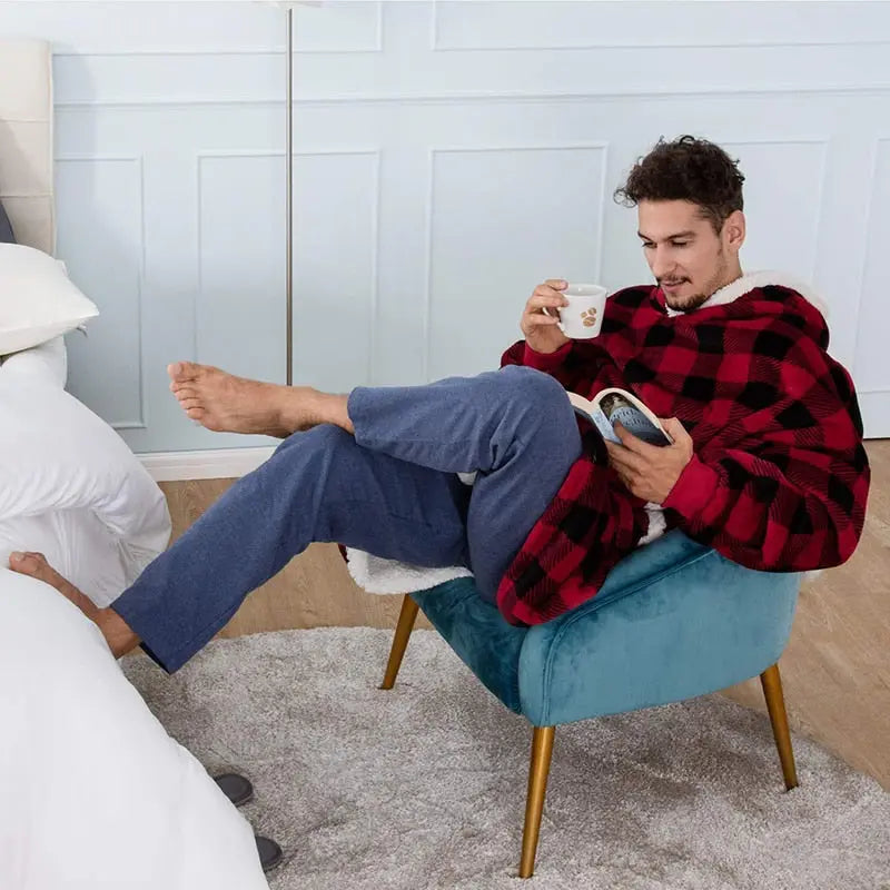 Person relaxing in red buffalo plaid Oversized Hoodie Blanket while reading and enjoying coffee in cozy teal armchair